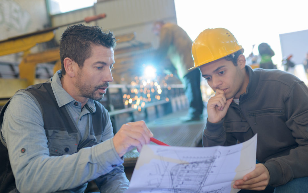 An engineer and client discuss design plans in an industrial manufacturing setting.