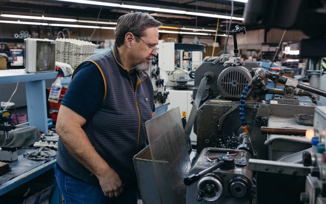 A precision grinder processes a machine spindle for additive manufacturing machines using OD grinding techniques. He is wearing a deep blue tshirt under a lighter blue vest.